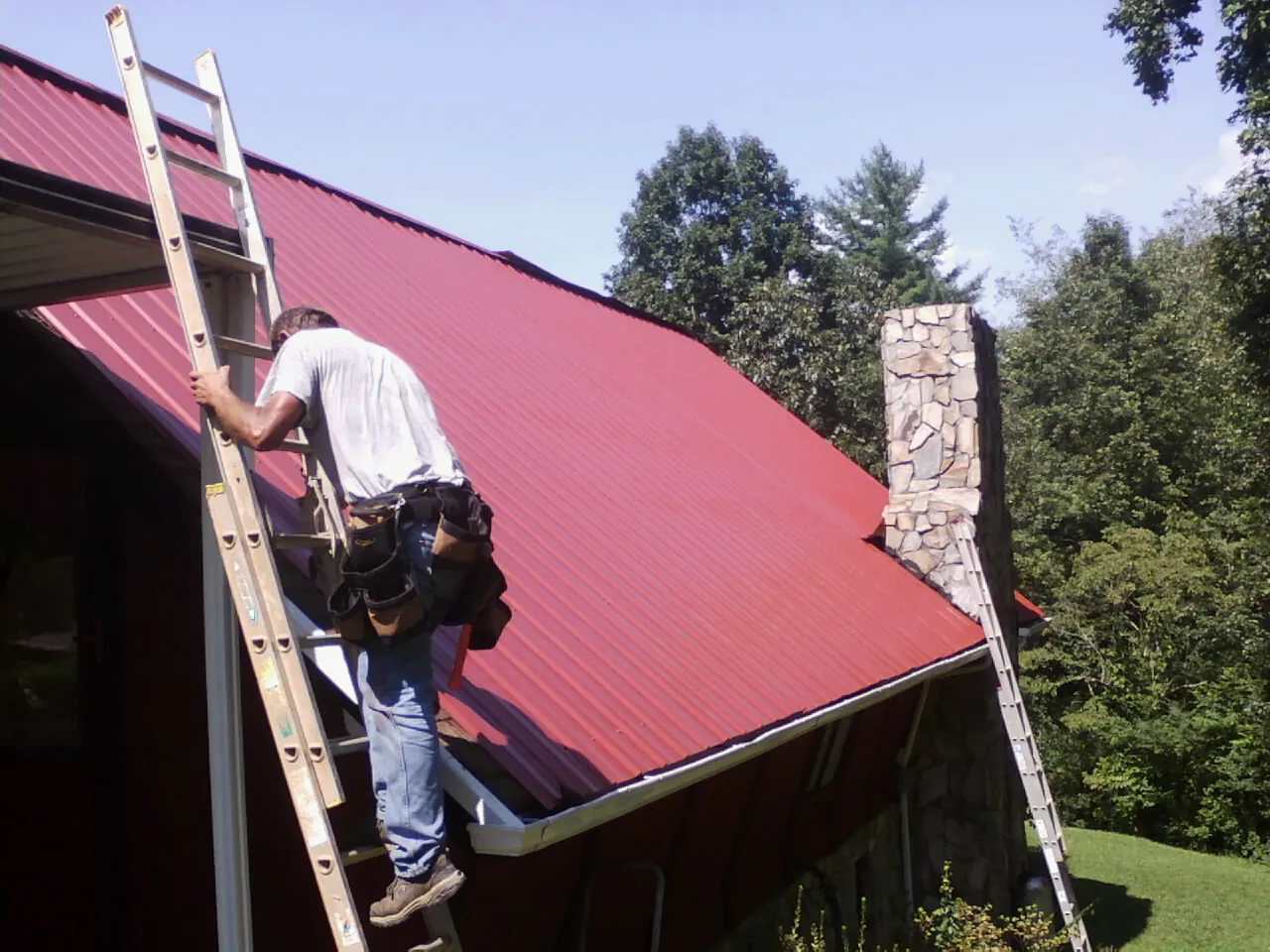 Roofer on a ladder beside a red metal roof and stone chimney in Wadesboro